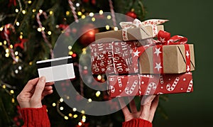 Hands of a woman in a red sweater holding a credit card and a gift box on the background of a Christmas tree close-up. Christmas