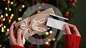 Hands of a woman in a red sweater holding a credit card and a gift box on the background of a Christmas tree close-up. Christmas