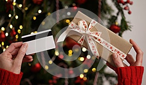 Hands of a woman in a red sweater holding a credit card and a gift box on the background of a Christmas tree close-up. Christmas