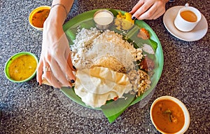 Hands of woman on table with traditional South Indian food thali with rice and spicy vegetables on palm leaf.