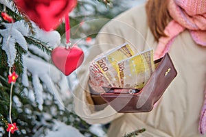 The hands of a woman in winter mittens hold a purse with money at the Christmas tree in a snow park on New Year`s Eve