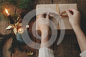 Hands wrapping stylish christmas gift in craft paper on rustic wooden background with candle, scissors, fir branches. Modern