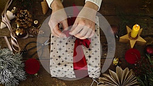 Hands wrapping stylish christmas gift with red ribbon on rustic wooden table with festive holiday decorations, top view. Happy Hol