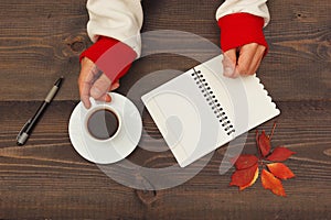 Hands of writer hold a cup of coffee at a wooden table with notebook and pen and autumn leaves