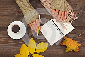 Hands of writer with a pen and notebook at a wooden table with cup of espresso and autumn leaves and scarf