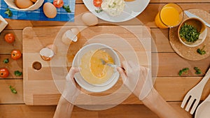 Hands of young Asian woman chef whisking egg into ceramic bowl cook omelette with vegetables on wooden board on kitchen table in