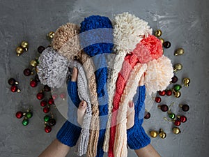 Hands of young beautiful woman in blue mittens holding many colored bright warm winter hats. On a gray background are bright