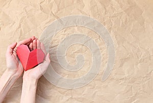 Hands of a young caucasian woman holding a folded paper red heart on a background of rough crumpled kraft