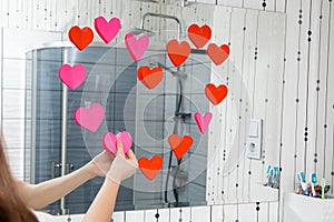 Hands of young girlwoman in the bathroom glue valentines red hearts on mirror in bathroom. Valentine day concept