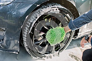 Hands of young man in jeans shirt holding green sponge, washing car wheel with foam. Cleaning of modern rims of luxury