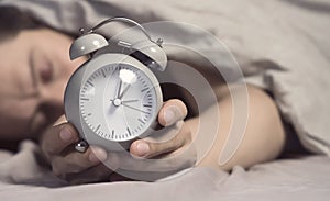 Hands of a young man from under the blankets hold an alarm clock in gray.