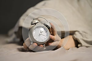 Hands of a young man from under the blankets hold an alarm clock in gray.