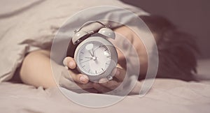 Hands of a young man from under the blankets hold an alarm clock in gray.