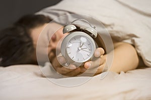 Hands of a young man from under the blankets hold an alarm clock in gray.