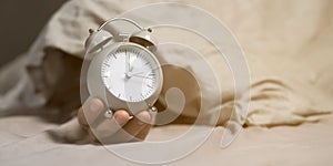 Hands of a young man from under the blankets hold an alarm clock in gray.