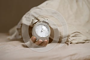 Hands of a young man from under the blankets hold an alarm clock in gray.