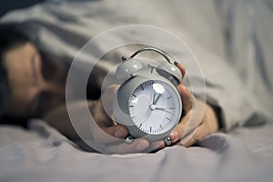 Hands of a young man from under the blankets hold a retro vintage alarm clock.