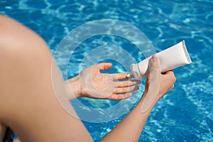 Hands of a young woman applying cream from a white tube of sunscreen on a background of blue water in a swimming pool. The concept