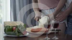 Hands young woman in jeans shorts cutting cabbage preparing romantic dinner. Her husband hugging her from behind. Glass