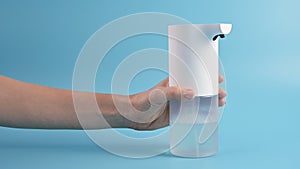 Hands of young woman picking up an automatic soap dispenser on blue background. Concept of hand washing, hygiene, health