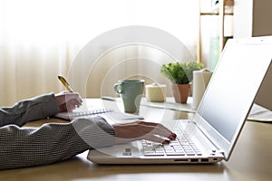 Hands of a young woman teleworking sitting at a desk writing in papers and typing on her laptop in a home office with a cup of