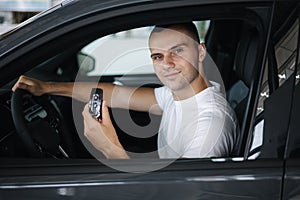 Handseome man in white t-shirt sitting in his new car in car showroom. Male hold car keys