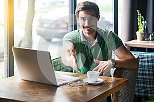 Handshake. Young happy businessman in green t-shirt sitting with laptop, toothy smile looking at camera and giving hand to