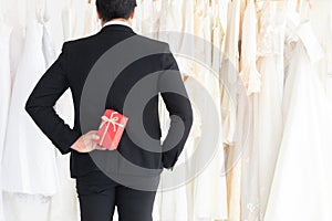 Handsom mix race groom hide the red gift box behind surprise his coupl bride in Wedding dress fitting toom, man in tuxedo black