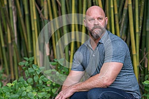 Handsome adult male model with a beard and bald head posing in a bamboo garden scene