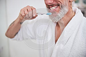 Handsome adult male in plush bathrobe brushing hos teeth in bathroom