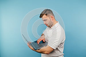 Handsome adult man with bristles enjoys laptop on blue background with serious face wearing white t-shirt. Isolated