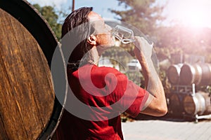 A handsome adult man stands near a wine barrel on a farm and tastes a glass of white wine.