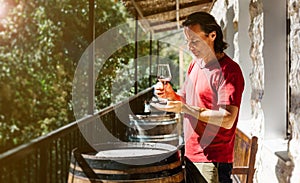A handsome adult man stands near a wine barrel on a farm and tastes a glass of white wine.