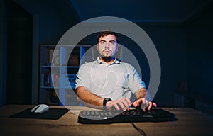 Handsome adult man in a white T-shirt sits at night at the computer and works with a serious face in a dark kiman with blue light