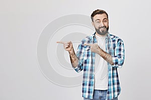 Handsome adult spouse with beard and moustache, smiling and expressing cheer while pointing left with two hands