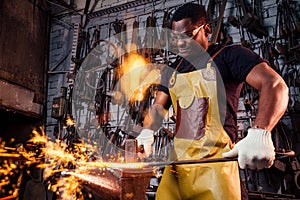 Handsome african americam man forging steel next to furnace in dark workshop. small business comcept