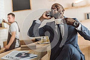 handsome african american businessman with cup of coffee laughing and talking on smartphone