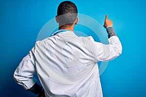 Handsome african american doctor man wearing coat and stethoscope over blue background Posing backwards pointing ahead with finger