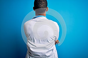 Handsome african american doctor man wearing coat and stethoscope over blue background standing backwards looking away with