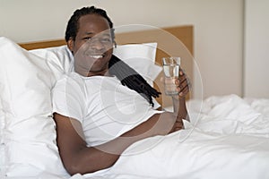 Handsome African American Guy Holding Glass With Water While Relaxing In Bed