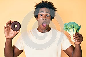 Handsome african american man with afro hair holding broccoli and chocolate donut celebrating crazy and amazed for success with