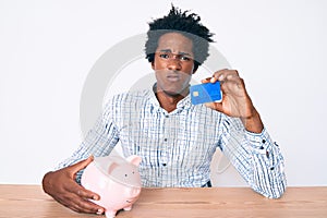 Handsome african american man with afro hair holding credit card and piggy bank skeptic and nervous, frowning upset because of