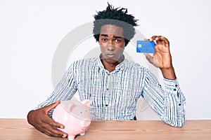 Handsome african american man with afro hair holding credit card and piggy bank skeptic and nervous, frowning upset because of