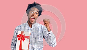 Handsome african american man with afro hair holding gift screaming proud, celebrating victory and success very excited with