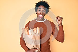 Handsome african american man with afro hair holding paper bag with bread screaming proud, celebrating victory and success very