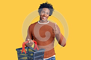 Handsome african american man with afro hair holding supermarket shopping basket smiling happy and positive, thumb up doing