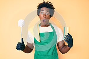 Handsome african american man with afro hair wearing apron holding scourer and toilet brush afraid and shocked with surprise and