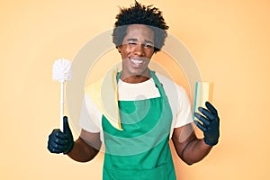 Handsome african american man with afro hair wearing apron holding scourer and toilet brush winking looking at the camera with