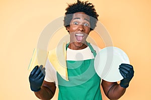 Handsome african american man with afro hair wearing apron holding scourer washing dishes celebrating crazy and amazed for success