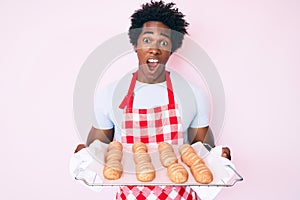 Handsome african american man with afro hair wearing baker uniform holding homemade bread celebrating crazy and amazed for success